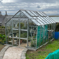 A glass Rhino Greenhouse stands on a grassy hill overlooking water. Inside, plants and gardening tools are visible. A gravel path leads to the entrance, and nearby are some outdoor structures.