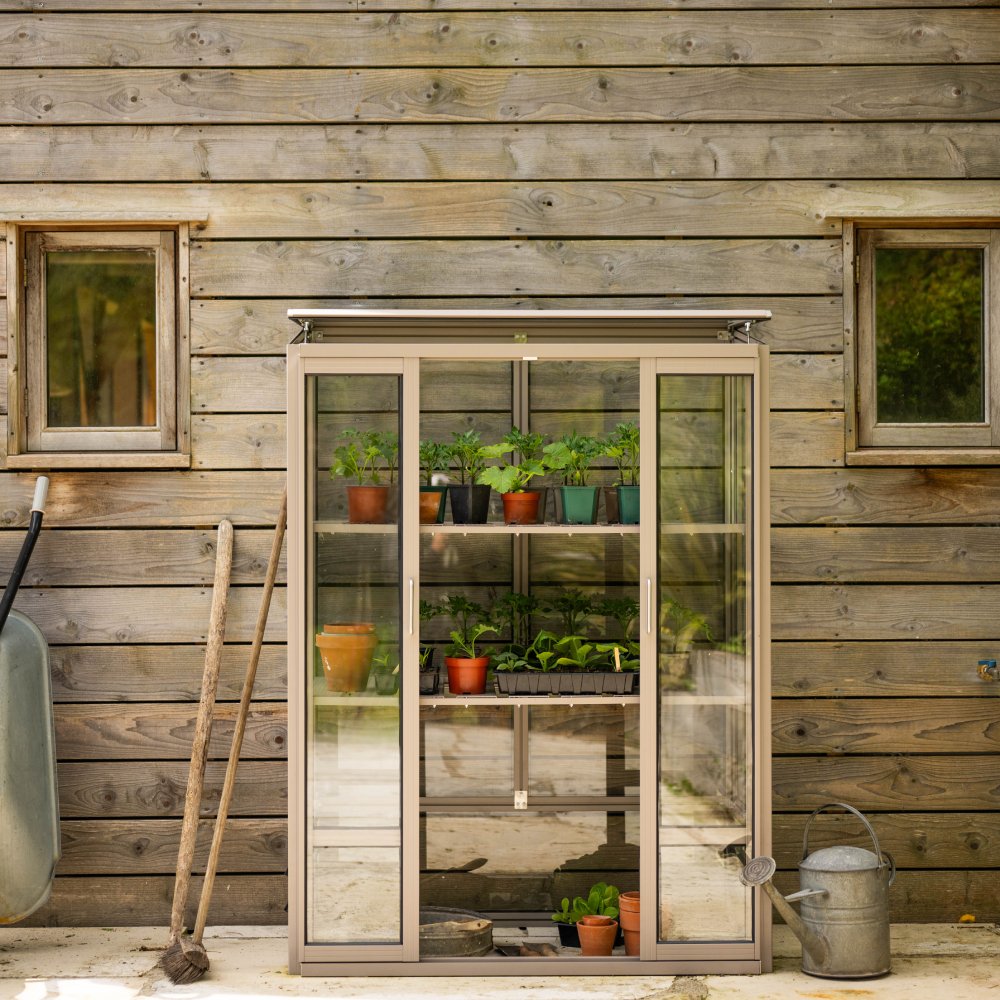 A small Rhino Greenhouse houses potted plants on shelves, standing against a wooden wall. Nearby, a wheelbarrow, rake, and watering can rest on the ground, conveying a gardening scene.