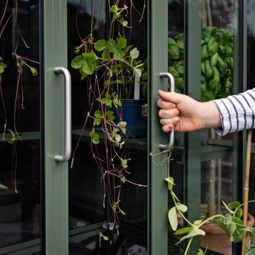 A hand grips a metallic handle, opening a green glass door adorned with trailing plants. Inside the Rhino Greenhouse, potted greenery and hanging vines enhance the lush, vibrant setting.