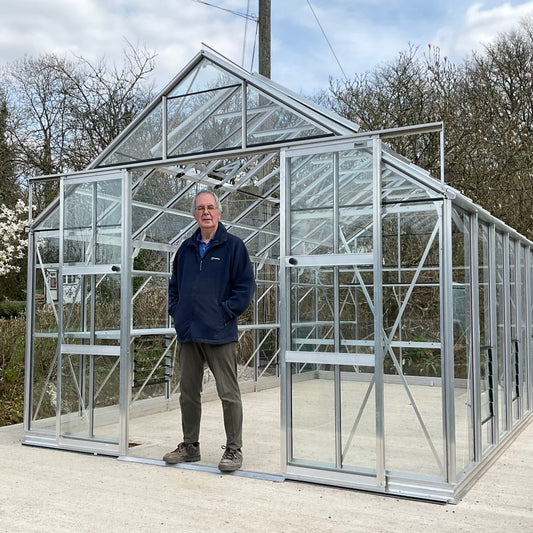 A man stands inside a large, glass-paneled Rhino Greenhouse with a sturdy metal frame. It is situated outdoors with trees and overcast sky in the background.