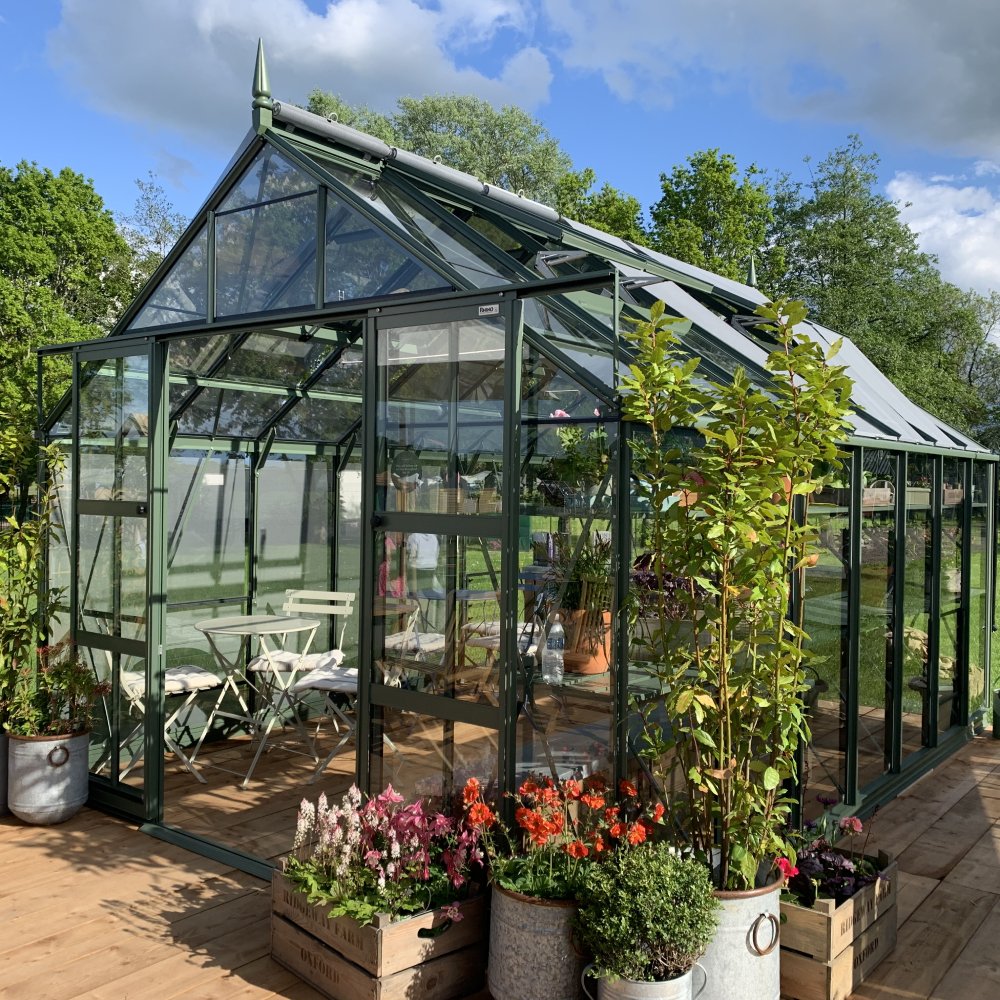 A green glass Rhino Greenhouse, housing potted plants and garden furniture, stands on a wooden deck. Surrounding it are lush green trees under a partly cloudy sky. Text: RHINO.