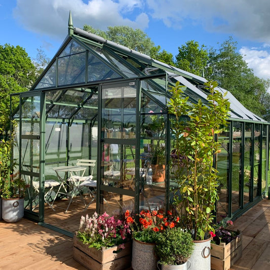 A green glass Rhino Greenhouse, housing potted plants and garden furniture, stands on a wooden deck. Surrounding it are lush green trees under a partly cloudy sky. Text: RHINO.