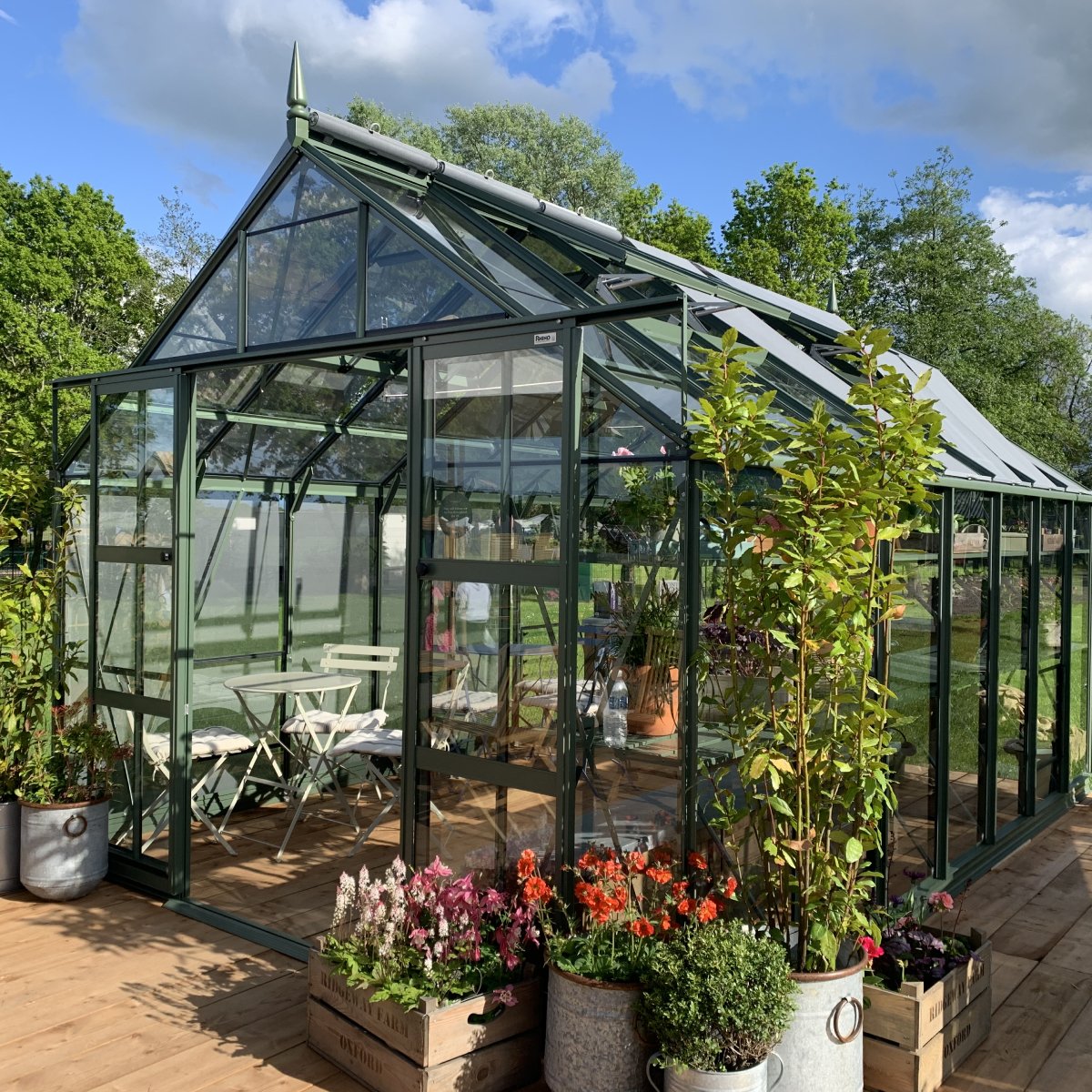 A glass Rhino Greenhouse stands on a wooden deck, surrounded by vibrant potted plants and outdoor furniture. The structure is set in a lush garden under a partly cloudy blue sky.