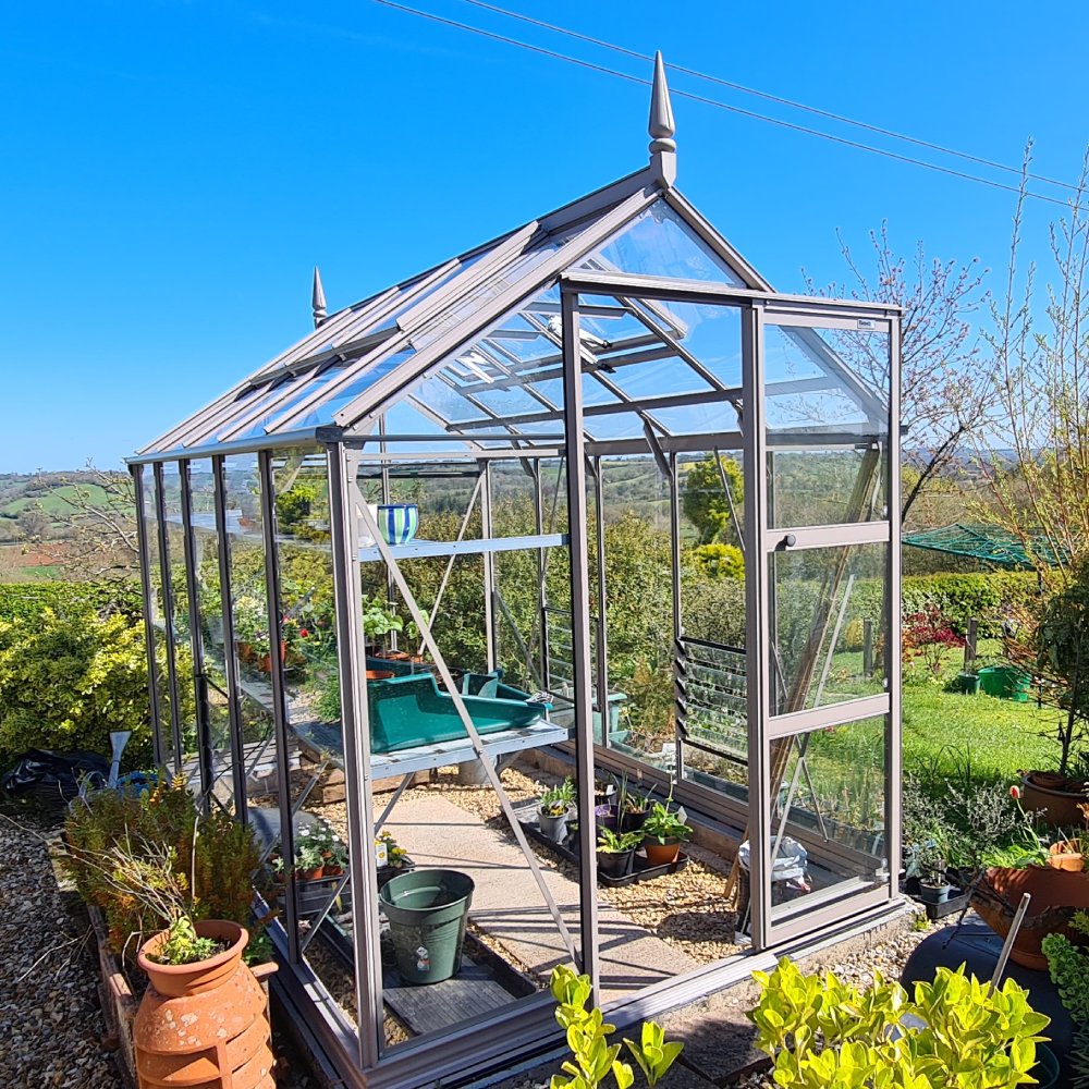 A glass Rhino Greenhouse stands in a garden, brimming with potted plants inside. It's surrounded by greenery and overlooks a distant landscape under a clear blue sky.