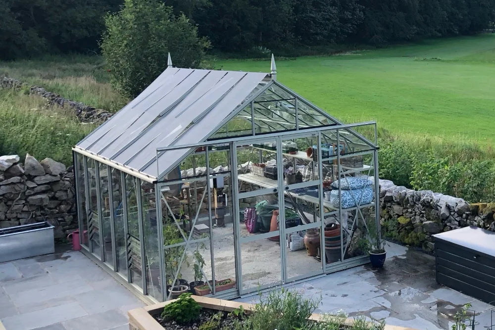 A glass Rhino Greenhouse stands on a paved area, housing various pots and garden supplies. It is surrounded by a stone wall, with a lush, green field and trees nearby.