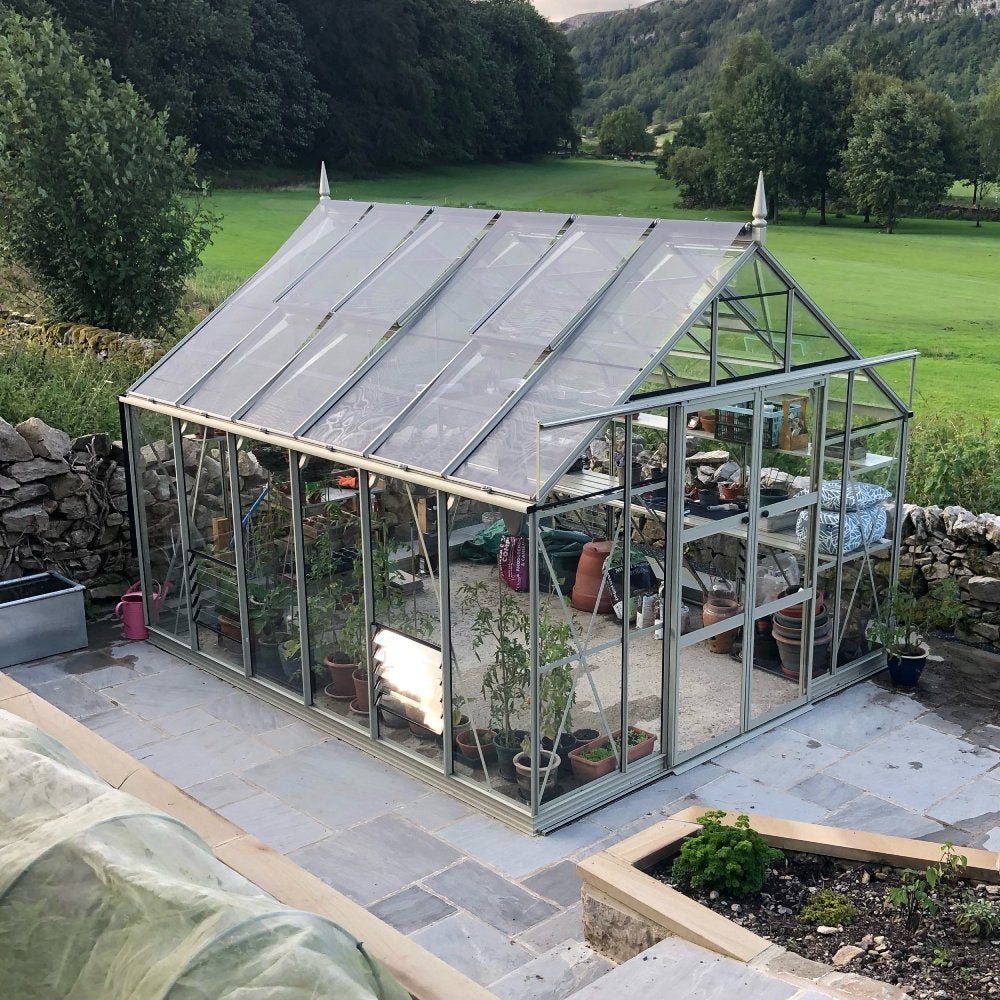 A Rhino Greenhouse with glass panels houses various potted plants on shelves, set on a stone patio surrounded by greenery and a lush landscape.