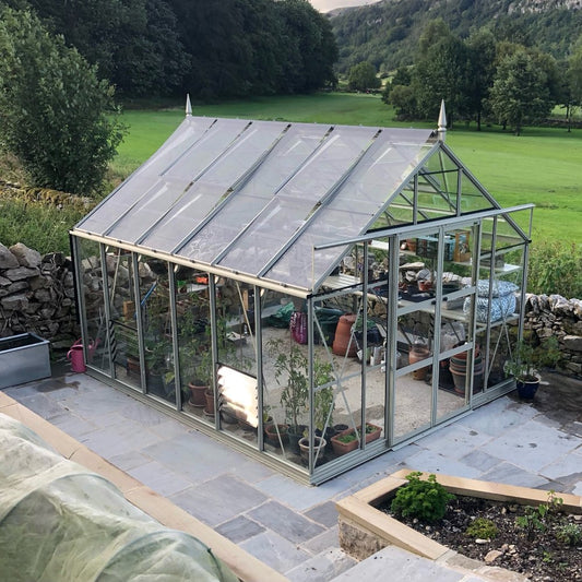 A Rhino Greenhouse with glass panels houses various potted plants on shelves, set on a stone patio surrounded by greenery and a lush landscape.