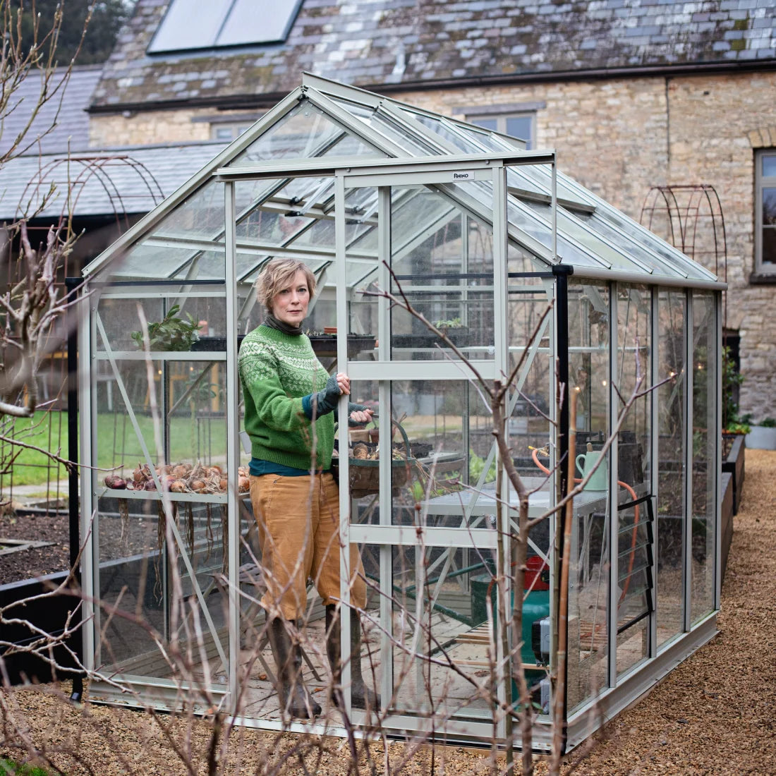 A person is standing inside a glass Rhino Greenhouse, sliding its door open. The Rhino Greenhouse is situated in a garden with a stone building in the background. The text Rhino is visible.