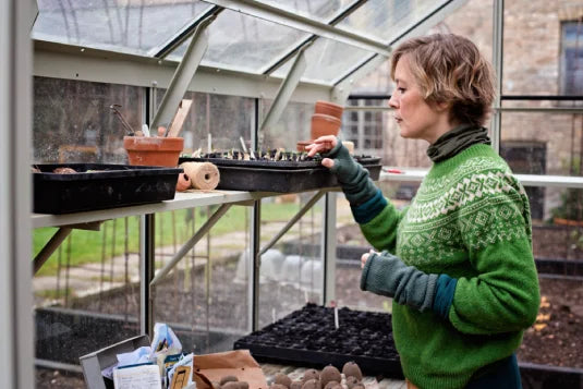 A person in a green sweater tends to seedlings in trays inside a Rhino Greenhouse with a view of a garden and a stone building outside. Gardening tools are visible nearby.