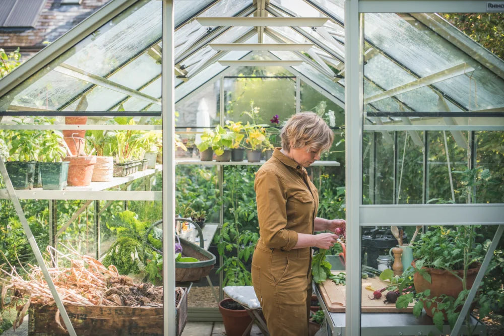 A person in a brown jumpsuit tends to plants inside a glass Rhino Greenhouse filled with potted greenery and gardening tools. Sunlight filters through the clear roof, illuminating the space.