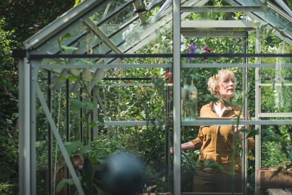 A person stands inside a glass Rhino Greenhouse, tending to plants surrounded by lush greenery. Sunlight filters through the transparent roof, creating a bright and nurturing environment.