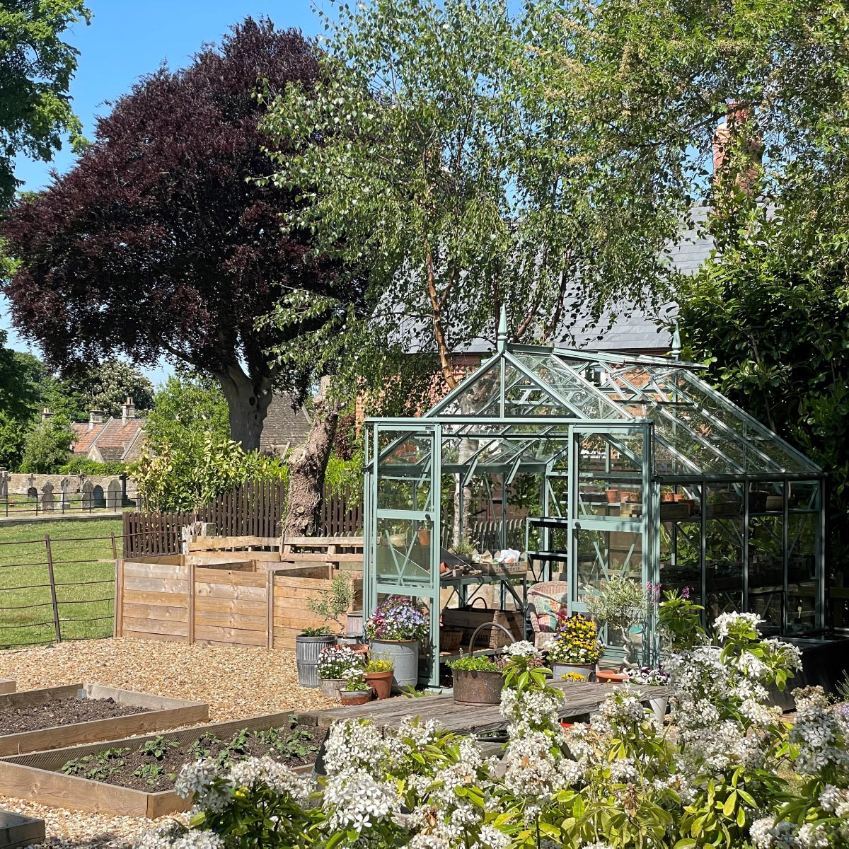 A glass Rhino Greenhouse stands amidst a lush garden, housing potted plants. Surrounded by trees and a wooden fence, it's set on gravel near Rhino metal raised garden beds and blooming flowers.