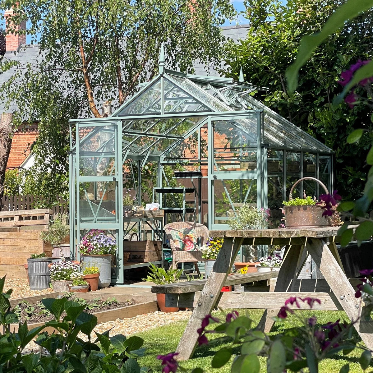 A glass Rhino Greenhouse stands open, surrounded by colorful potted plants and a wooden picnic table in a lush garden setting under a clear blue sky.