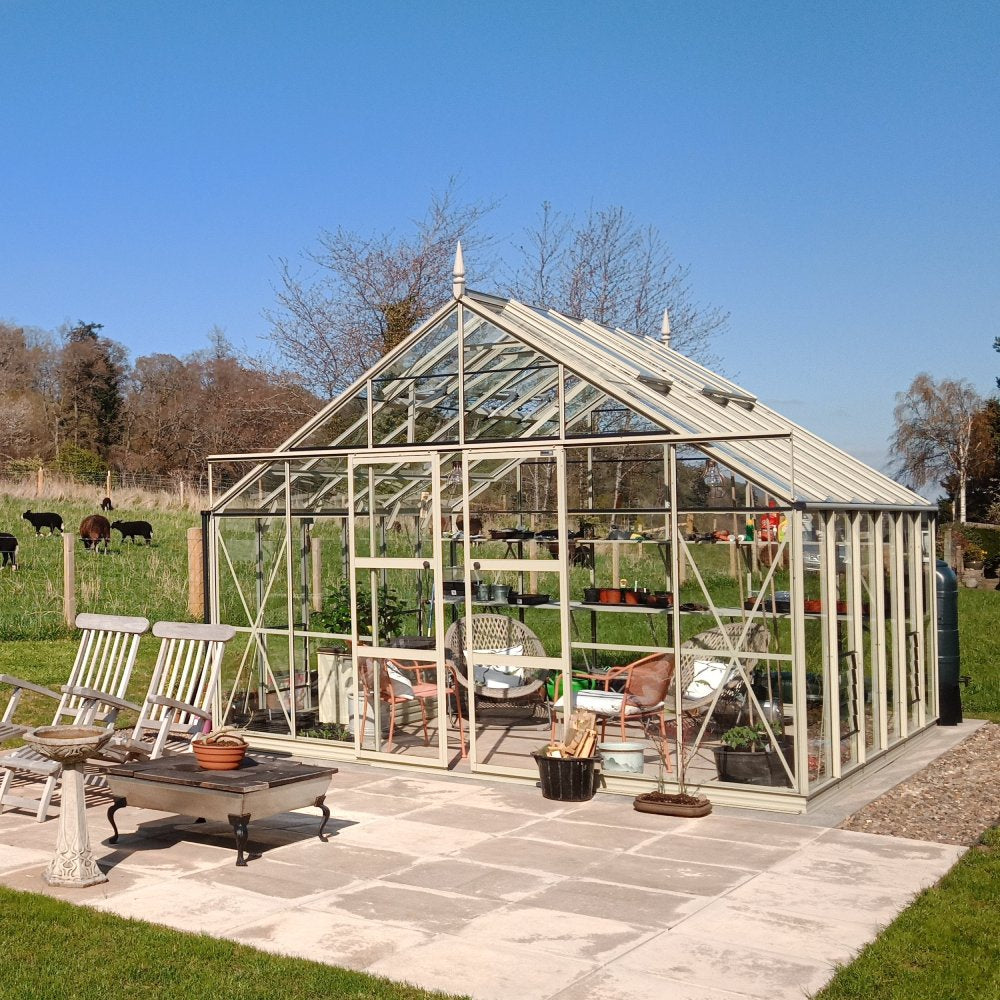 A Rhino Greenhouse stands on a tiled patio, housing plants and garden furniture. Surrounding the structure is a grassy field with grazing cows and distant trees under a clear blue sky.