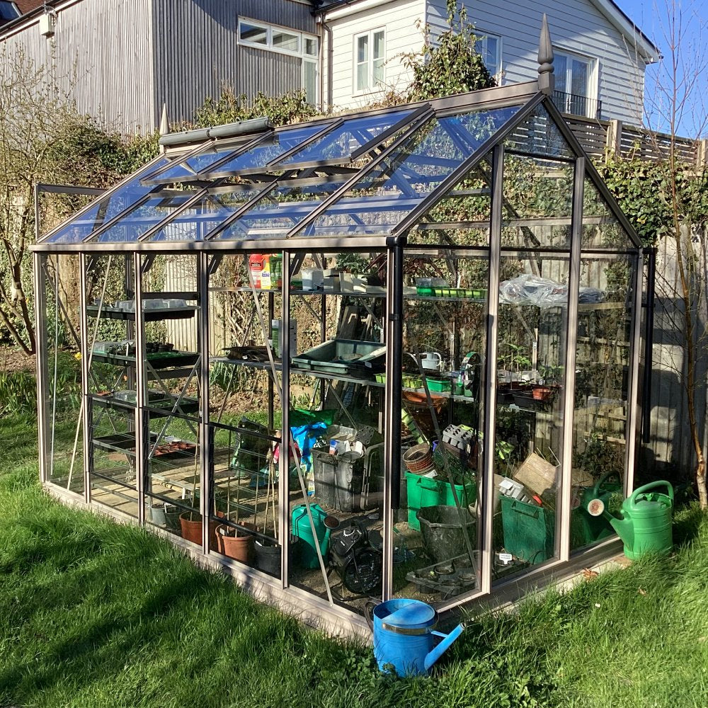 A glass Rhino Greenhouse full of gardening tools and pots stands in a grassy backyard with neighboring houses and greenery in the background, emphasizing outdoor gardening activities.