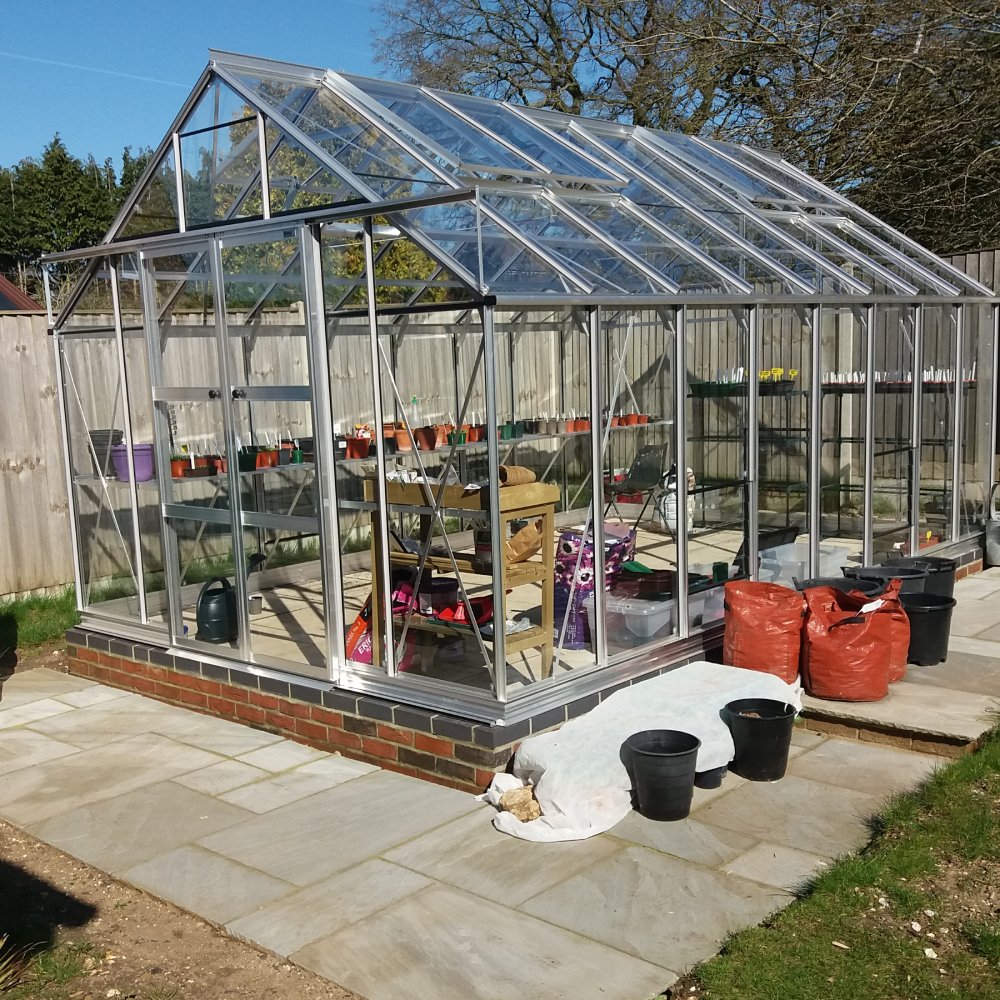 A clear glass Rhino Greenhouse stands on a brick base, containing various gardening tools and pots. It is surrounded by a stone patio and a wooden fence, under a blue sky.