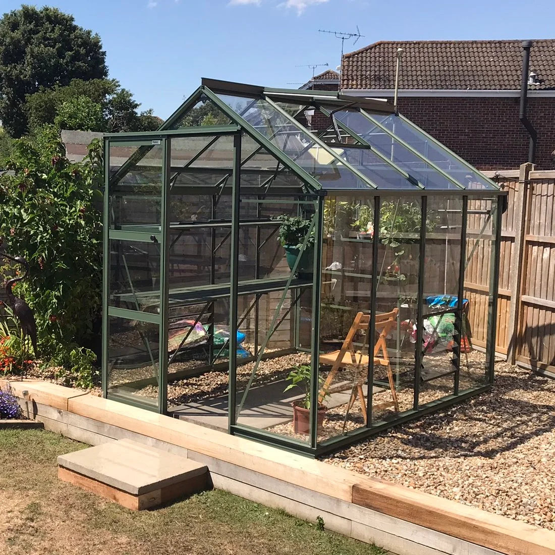 A glass Rhino Greenhouse sits in a garden, surrounded by gravel and wooden boards, with plants and gardening tools visible inside. Nearby, wooden fences and houses are in the background.