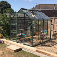 A glass Rhino Greenhouse sits in a garden, surrounded by gravel and wooden boards, with plants and gardening tools visible inside. Nearby, wooden fences and houses are in the background.