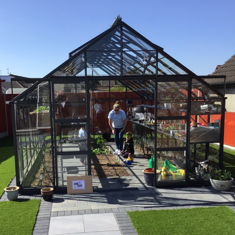 A glass Rhino Greenhouse houses a woman and child gardening. Pots and gardening supplies surround them on a paved area, with a vibrant yard and red fences in the background.