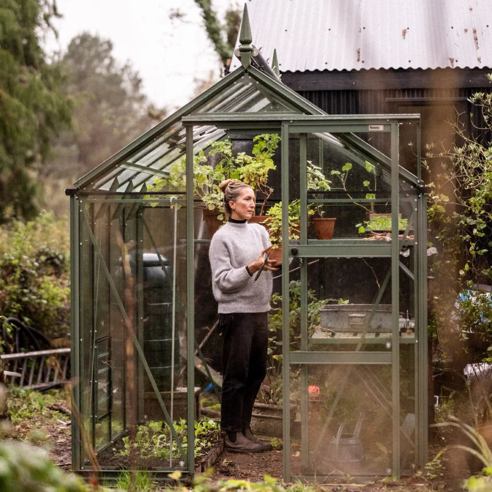A person tends potted plants inside a glass Rhino Greenhouse with green metal framing. The Rhino Greenhouse is situated in a lush garden area, next to a dark corrugated metal building.