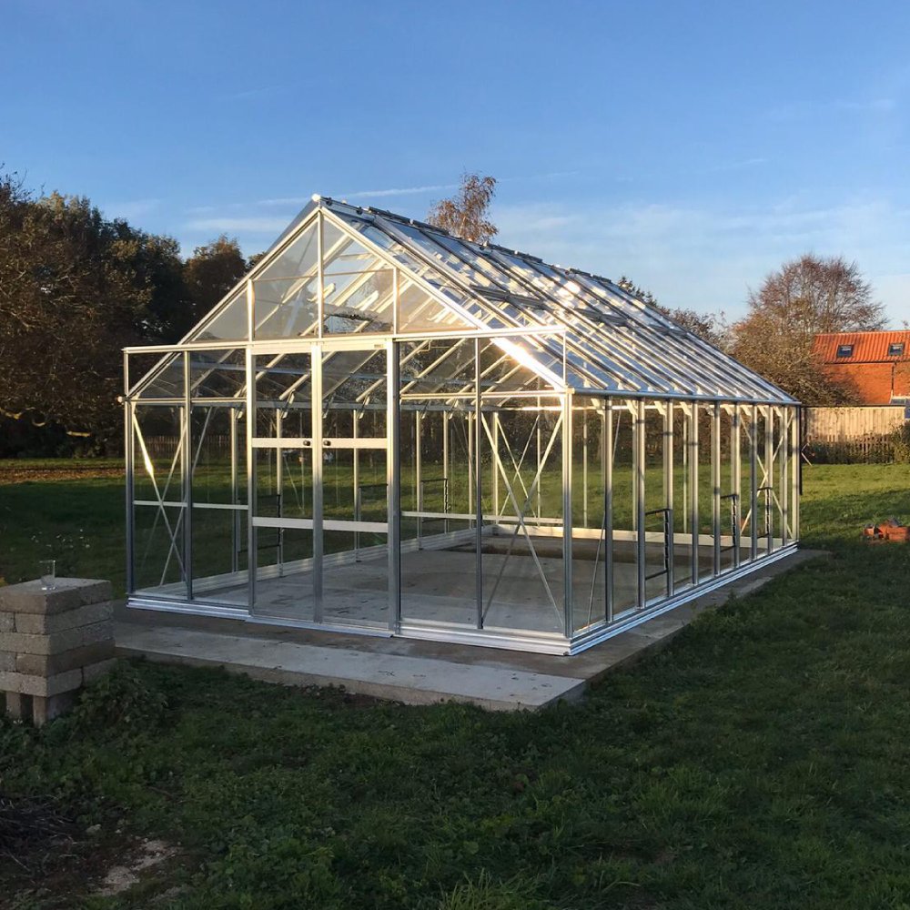 A large glass and metal Rhino Greenhouse stands empty on a concrete slab, surrounded by green grass and trees, with a clear blue sky in the background.