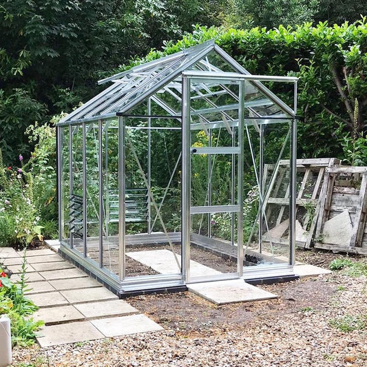 A glass Rhino Greenhouse stands on a paved area in a garden, surrounded by lush greenery and wooden pallets.