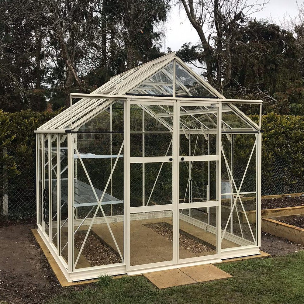 A glass and metal Rhino Greenhouse stands neatly on a wooden base, framed by trees and bushes, with visible interior plant beds and a sliding door for entry.