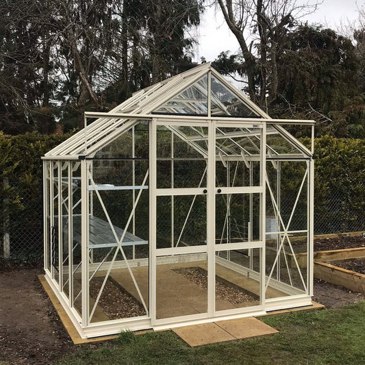 A glass and metal Rhino Greenhouse stands neatly on a wooden base, framed by trees and bushes, with visible interior plant beds and a sliding door for entry.