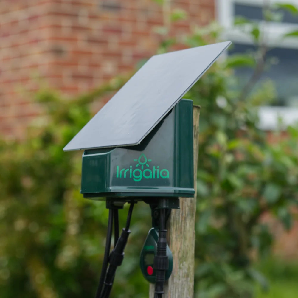 Solar-powered irrigation device mounted on a wooden post with Irrigatia label, surrounded by garden greenery and a blurred brick wall in the background.