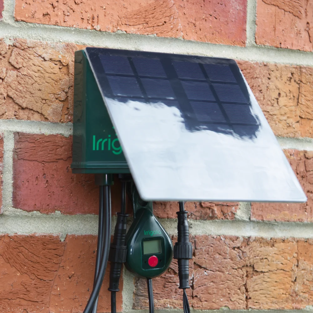 Solar-powered controller mounted on a brick wall operates irrigation systems. The unit features a solar panel and display, with the words Irri and Irrigatia visible on the device.