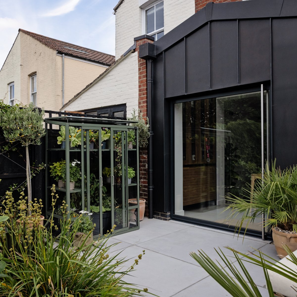 Rhino Greenhouse containing lush plants is positioned on a patio. It's adjacent to a modern building with black and brick exterior, surrounded by potted plants and a clear, blue sky above.