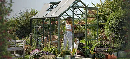 A glass and metal Rhino Greenhouse shelters a woman tending plants, with a dog nearby. Surrounding it are various potted flowers and garden tools, set in a lush garden.