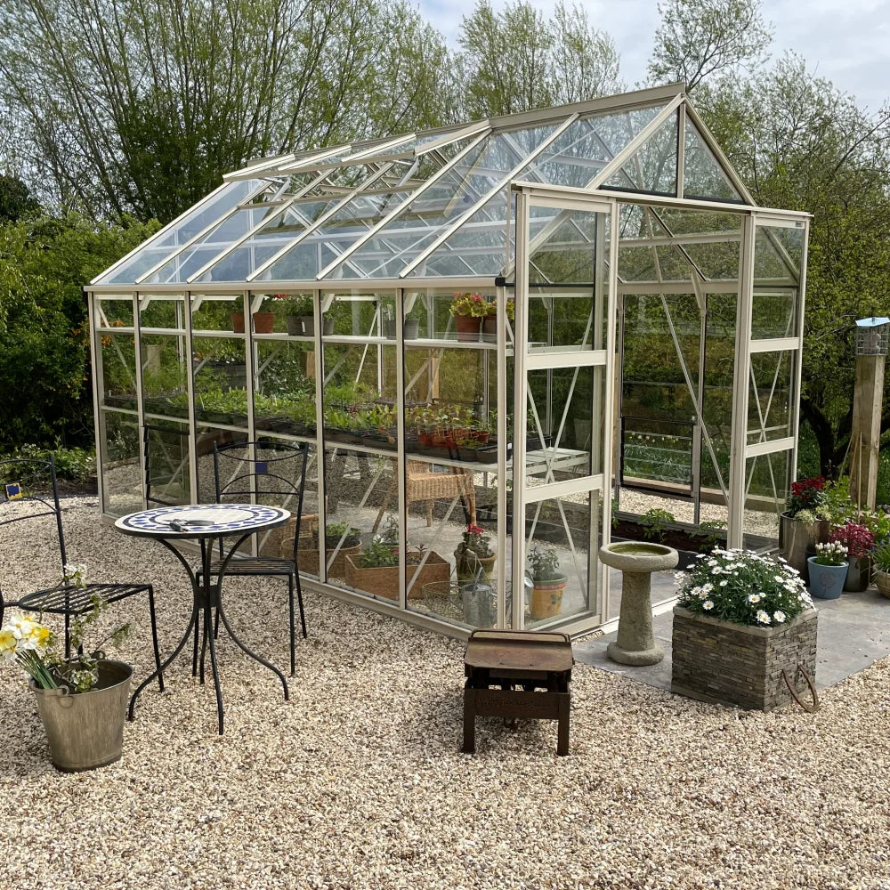 A glass Rhino Greenhouse houses potted plants, with a table, chairs, and decorative items nearby, set on gravel amidst lush, green trees.