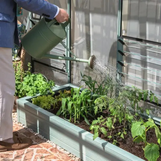 A person in a blue jacket waters plants with a green watering can in a Rhino metal raised garden bed next to a Rhino Greenhouse. Pebbled ground and nearby plants create a garden setting.