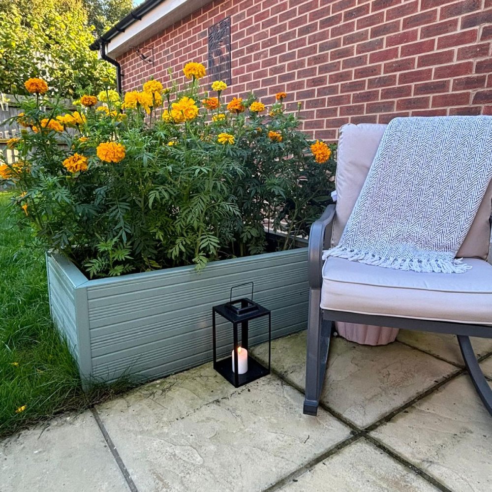 A planter with bright orange marigolds stands beside a cushioned chair draped with a patterned blanket. A lit lantern sits on a stone patio next to a brick wall.