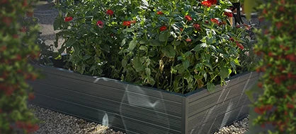A Rhino metal raised garden bed filled with lush green plants and red flowers stands on a gravel surface, framed by blurred foliage on both sides.