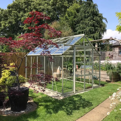 A glass-paneled Rhino Greenhouse stands on a grassy garden path, surrounded by various trees and plants, with a clear blue sky overhead.