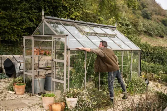 A man leans against a glass Rhino Greenhouse, inspecting plants inside. The Rhino Greenhouse is surrounded by potted plants and greenery, set in a lush garden with trees in the background.