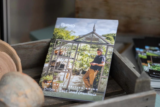 A brochure featuring Rhino Rhino Greenhouses is displayed in a wooden crate. The cover shows a person standing inside a Rhino Greenhouse. Text reads: THE GREAT BRITISH RHINO Britain's Best Loved Rhino Greenhouse.