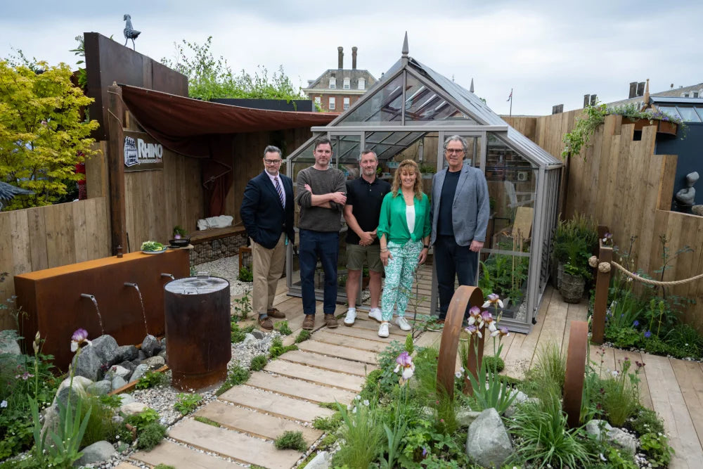Five people stand in front of a glass Rhino Greenhouse on a wooden deck surrounded by garden plants. A sign reads Rhino Rhino Greenhouses, with water features and decorative elements visible nearby.