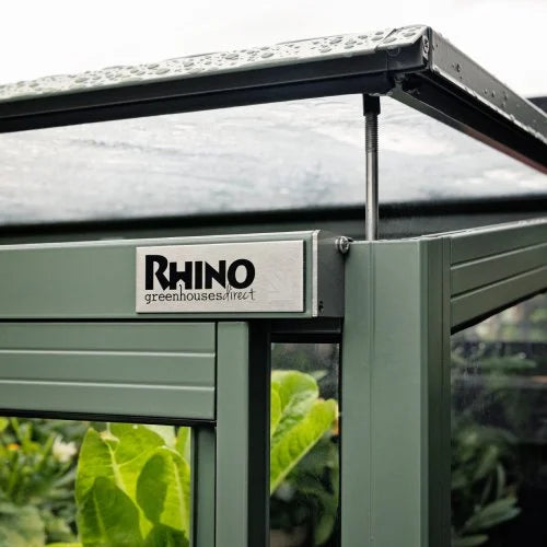 A green Rhino Rhino Greenhouse door slightly open, showing leafy plants inside. A metal plate reads RHINO Rhino Greenhouses direct. The surrounding area shows a garden environment.