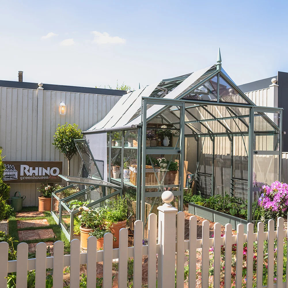 A glass Rhino Greenhouse, housing potted plants and flowers, stands in a garden setting. A sign reads Rhino Rhino Greenhouses direct. Enclosed by a white picket fence, it's bathed in sunlight.