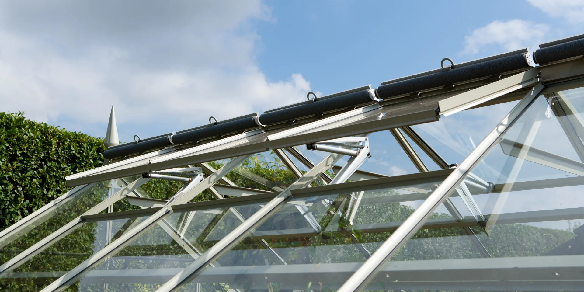 Rhino Greenhouse roof with open vents allowing airflow, surrounded by neatly trimmed hedges and a clear blue sky with clouds.