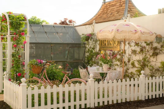 A quaint garden scene features a small Rhino Greenhouse surrounded by flowering plants. A pink bicycle rests against a white picket fence. A patio set and parasol sit under a sign reading RHINO Rhino GreenhouseS.