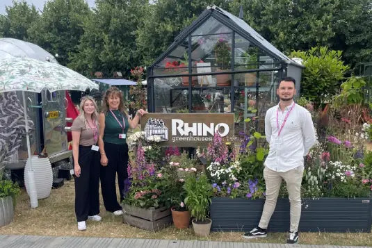 A glass Rhino Greenhouse displays various plants, surrounded by blooming flowers. Three people stand smiling beside a sign reading RHINO Rhino Greenhouses, set in a garden-like setting with trees and a trailer nearby.