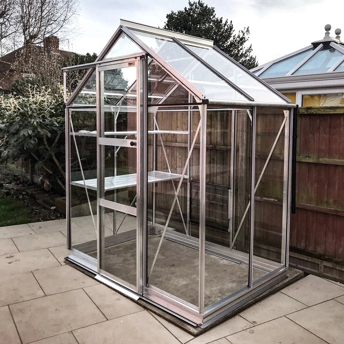 A small Rhino Greenhouse stands on a paved patio with a clear glass structure and metal frame. It is situated in a fenced backyard beside a larger conservatory building.