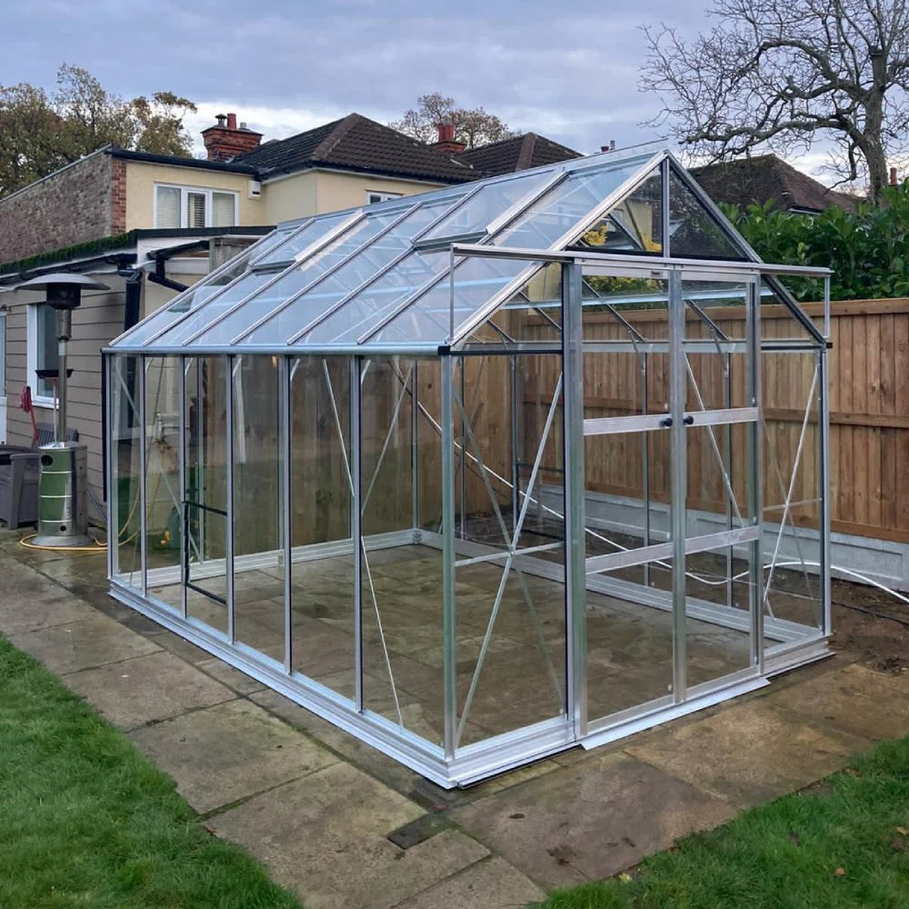 A glass and metal Rhino Greenhouse stands on a paved area next to a house, bordered by a wooden fence and garden with grass and trees.