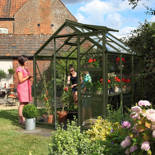 A green glass Rhino Greenhouse shelters flourishing plants and flowers. Two people work inside, while another stands outside smiling. It's set in a garden with a brick building nearby.