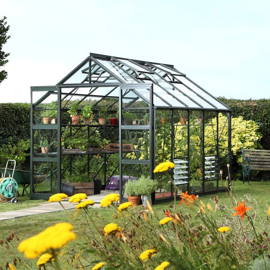 Rhino Greenhouse displaying potted plants on shelves, situated in a garden with colorful flowers and a hedge. Displayed text includes Rhino. A bench and garden tools are visible nearby.