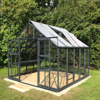A glass and metal Rhino Greenhouse stands empty on a concrete slab, surrounded by lush green foliage. Sunlight filters through its transparent panels, highlighting its sturdy, modern structure.